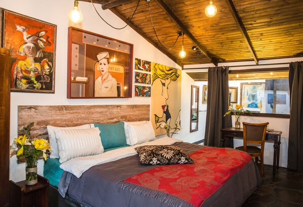 A bedroom with a wooden headboard, a bed with a red and grey bedspread, a wooden desk, and a chair. There are paintings on the walls and a vase with flowers on the nightstand. - LA COLINA Hotel Cottage (Bogotá)