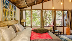 A bedroom with a bed, a nightstand, a lamp, a window with curtains, and a painting on the wall.