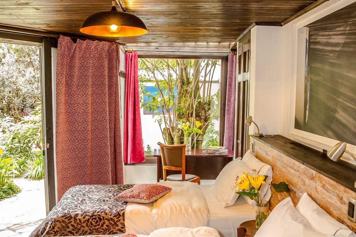 A bedroom with two beds, a window with curtains, a desk with a chair, and a vase with flowers.