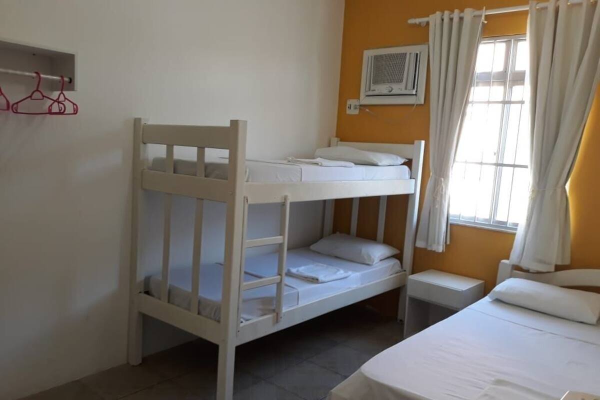 A room with a bunk bed, a single bed, a bedside table, a wall-mounted air conditioner, and a window with curtains.