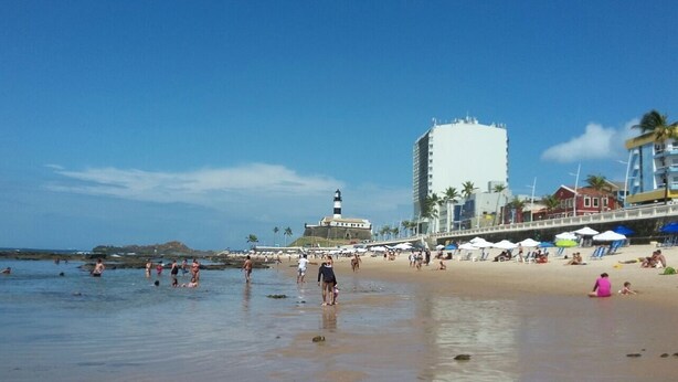 Una playa con gente, un faro y edificios al fondo.