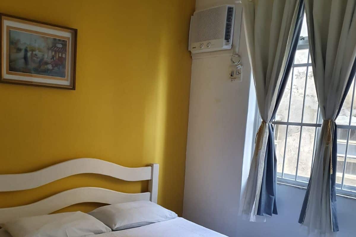 A hotel room with a bed, a painting on the wall, an air conditioning unit, and a window with curtains.