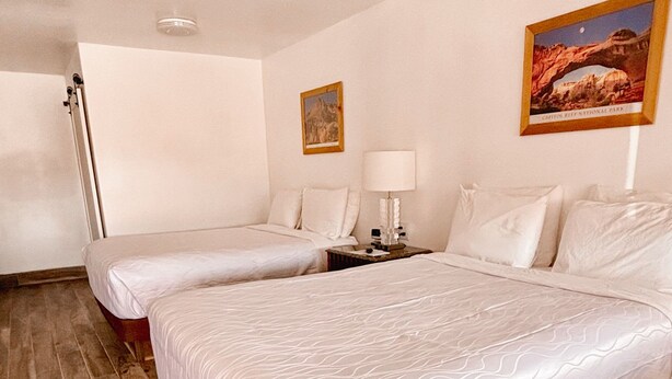A hotel room with two beds, a nightstand, a lamp, and a framed picture on the wall. - Whispering Sands Motel (Hanksville)