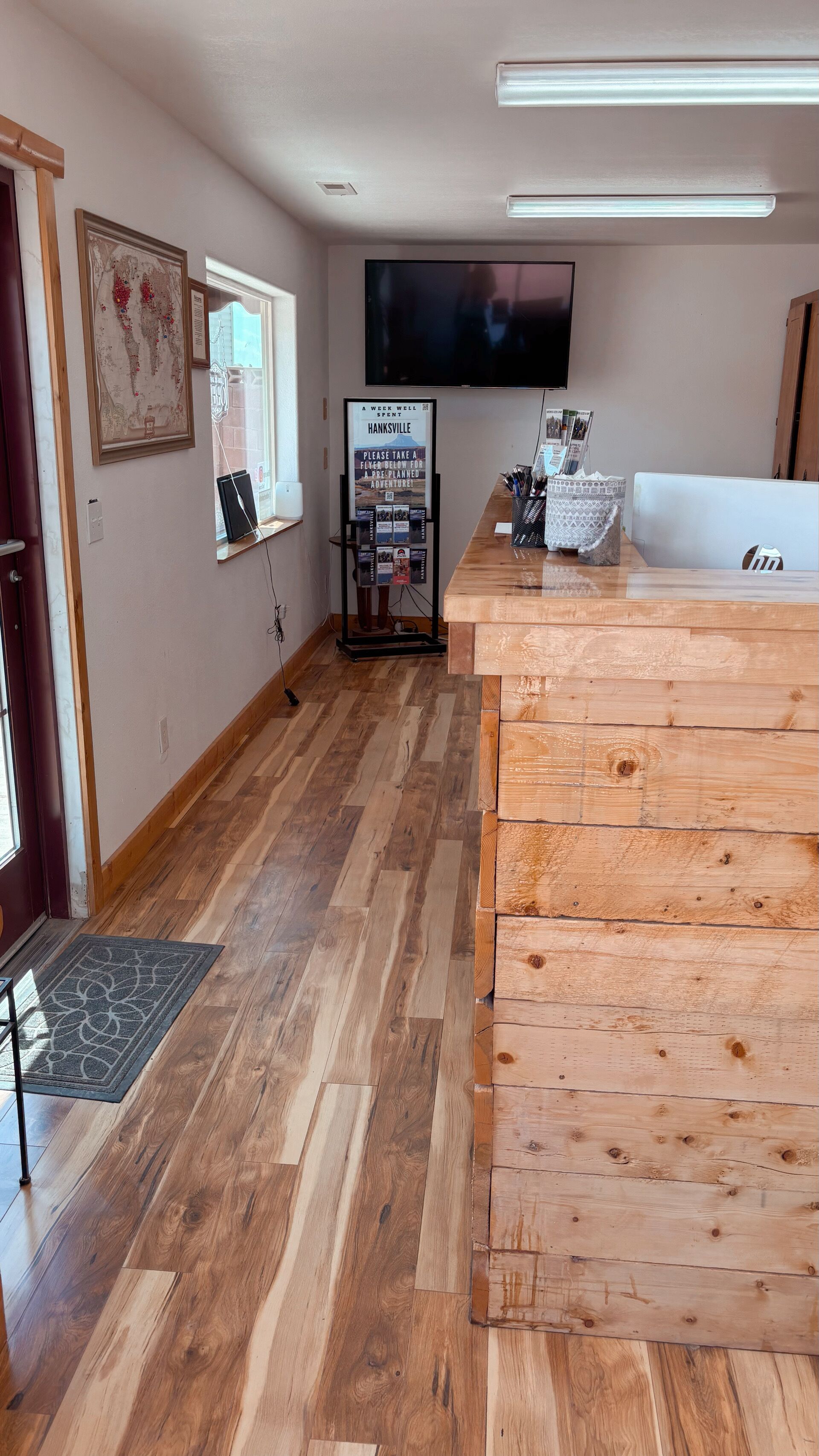 Un espace d’accueil d’hôtel avec un comptoir de réception en bois, un téléviseur à écran plat fixé au mur, une carte du monde encadrée et un panneau d’affichage avec un message.