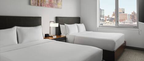 A hotel room with two beds, a nightstand with a lamp, a city view, and a piece of abstract art on the wall.