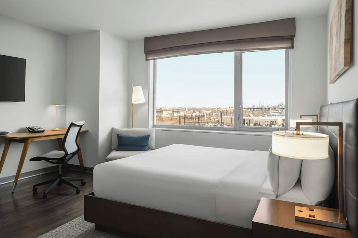 Habitación de hotel con cama, escritorio, silla, lámpara y vista a la ciudad.