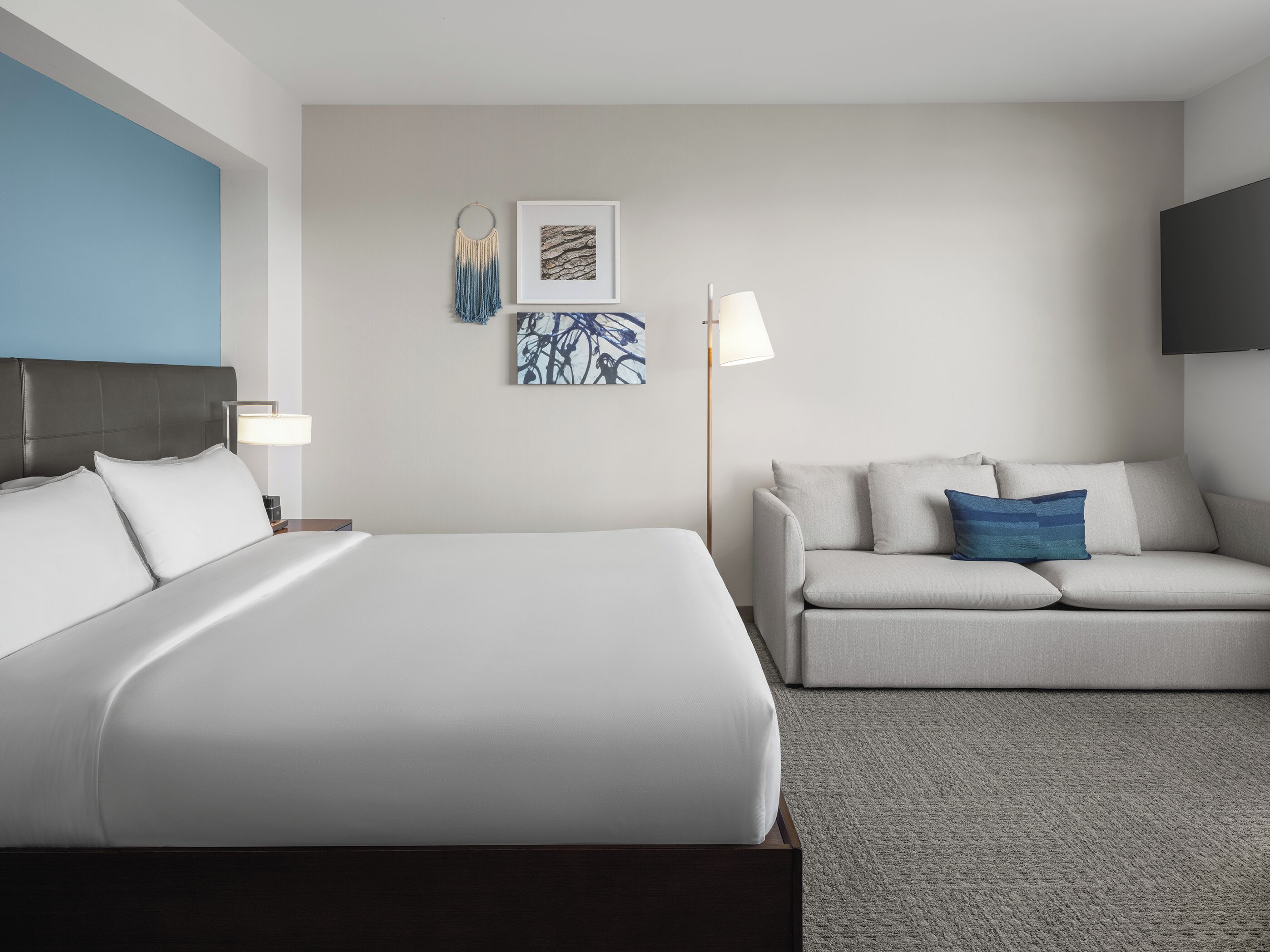 A hotel room with a bed, a sofa, a wall-mounted TV, and artwork.