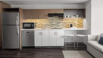 Una cocina moderna con un refrigerador de acero inoxidable, un microondas y un lavavajillas. Hay una encimera de madera con salpicadero de azulejos, y una pequeña zona de estar con un sofá y dos taburetes.