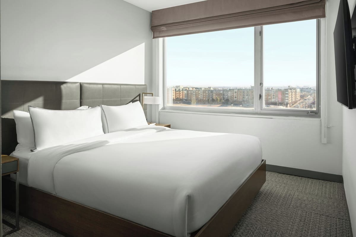 Habitación de hotel con una cama grande, una mesita de noche, una lámpara, una ventana con vistas a la ciudad y un televisor de pantalla plana.
