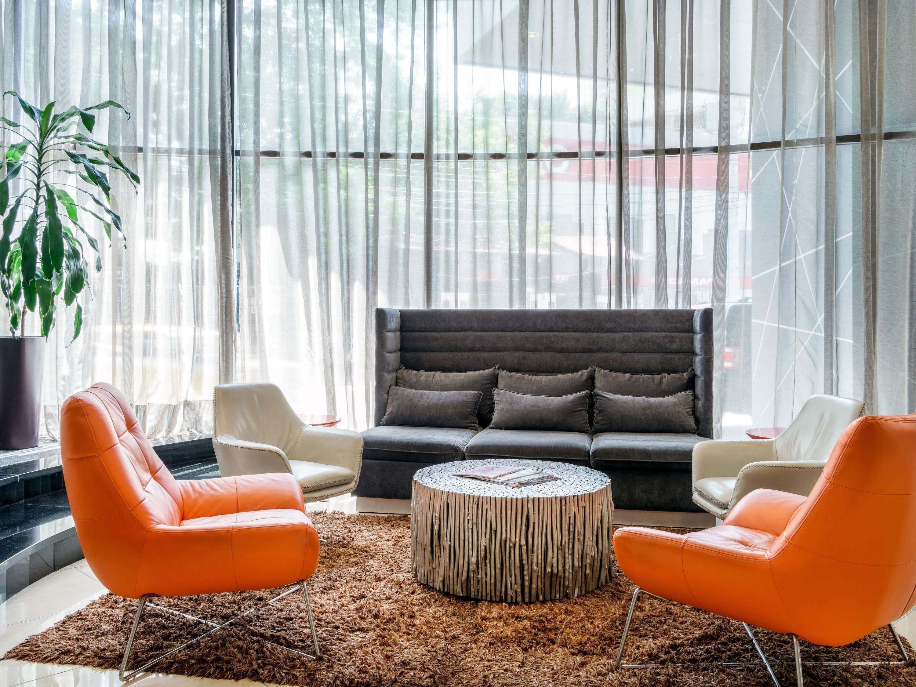A modern living room with a sofa, two orange chairs, a round coffee table, and a large window with sheer curtains.