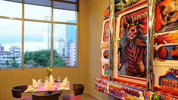 Un comedor con una mesa dispuesta para dos personas, una ventana con vista a la ciudad y una máquina recreativa de colores.