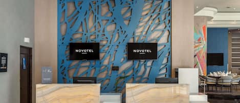 A hotel reception area with two reception desks, a blue decorative wall, and a large TV screen displaying 'NOVOTEL PANAMA CITY'.