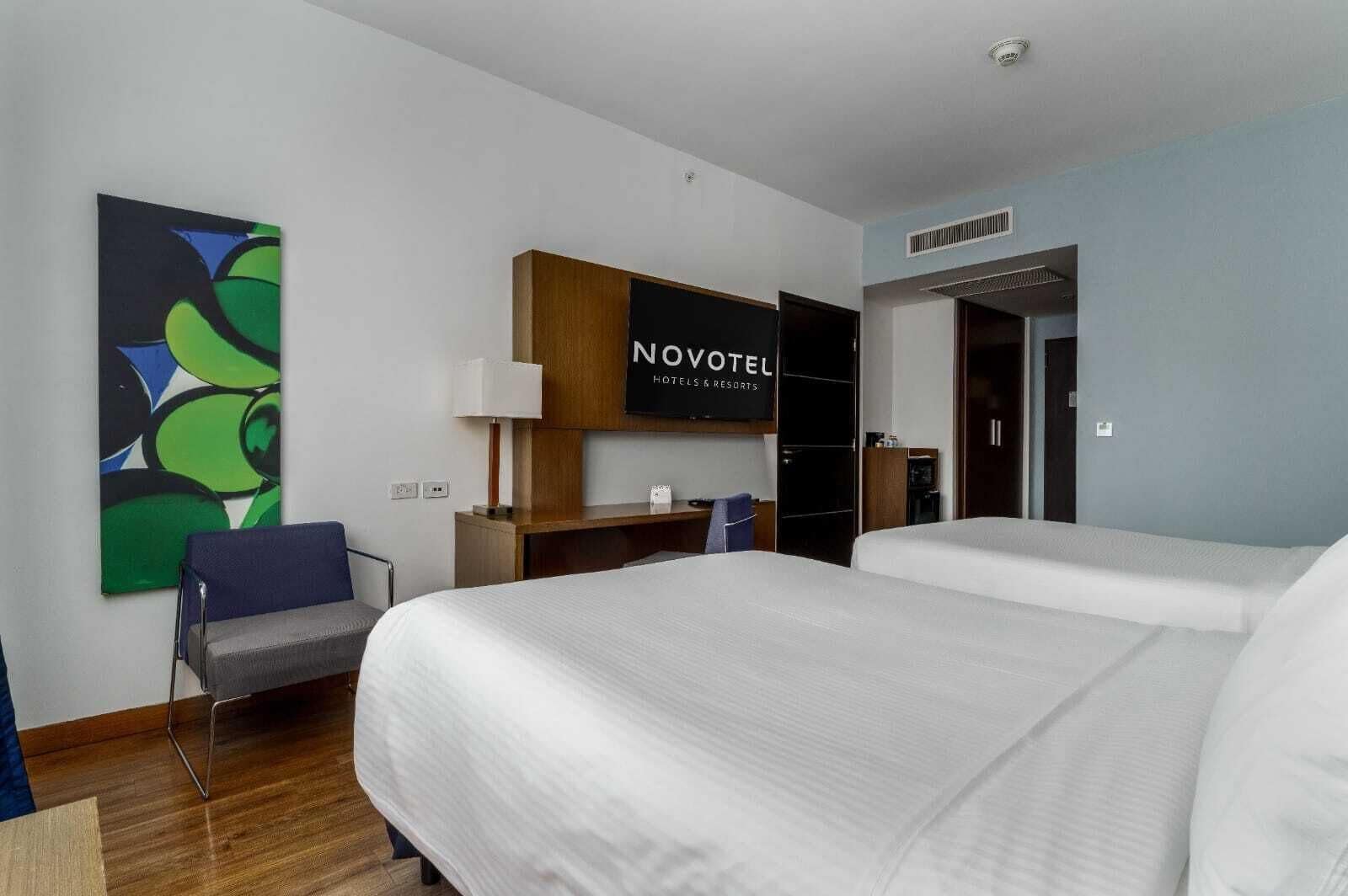 A hotel room with two beds, a desk, a chair, a TV, and a painting on the wall.