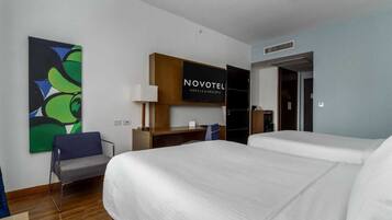 Quarto de hotel com duas camas, uma escrivaninha, uma cadeira, uma televisão e um quadro na parede.
