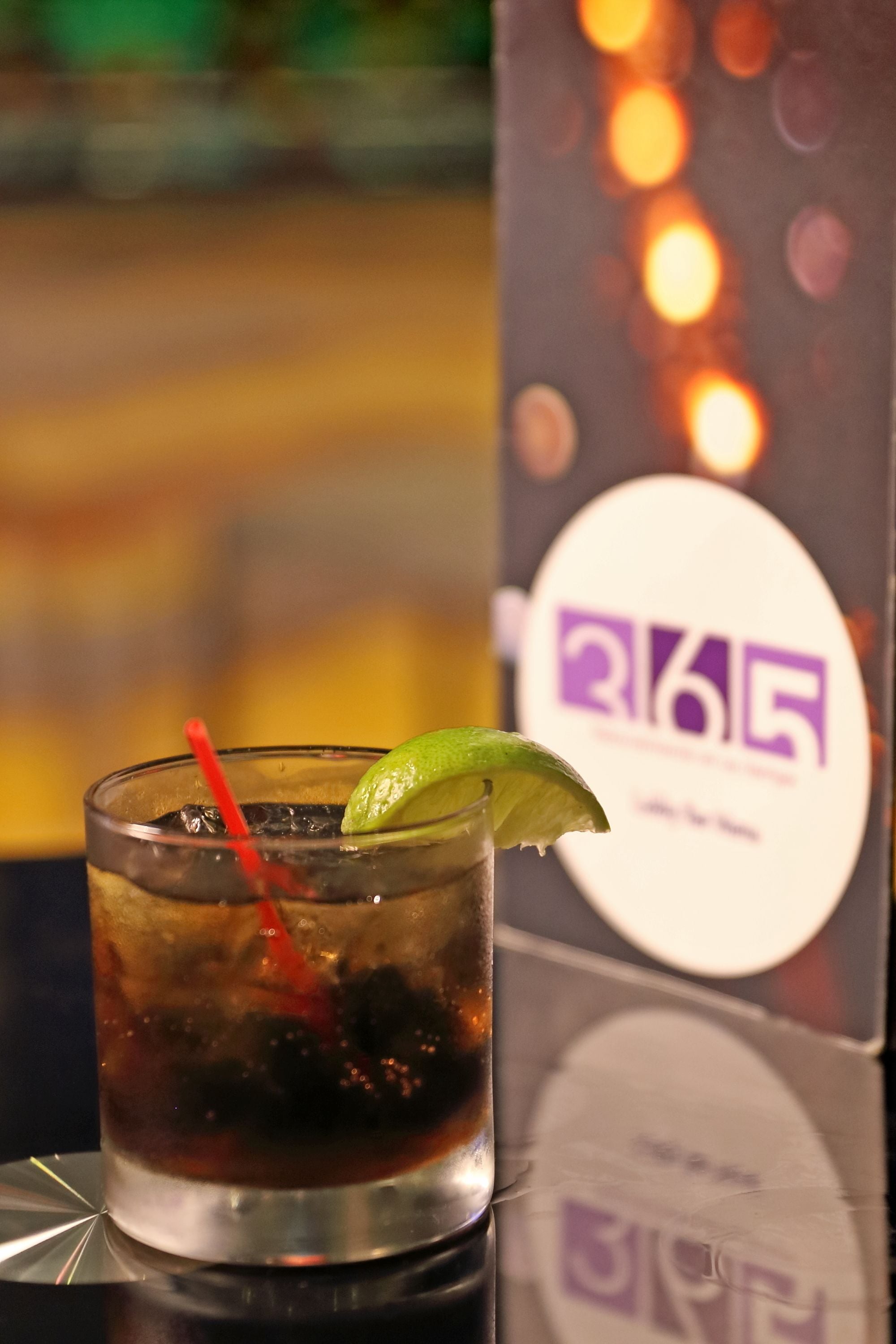 A cocktail glass with a lime wedge and a straw, placed on a reflective surface with a blurred background featuring a sign with the number "365".