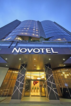 A modern hotel entrance with a prominent 'NOVOTEL' sign, large glass windows, and geometric light patterns on the facade.