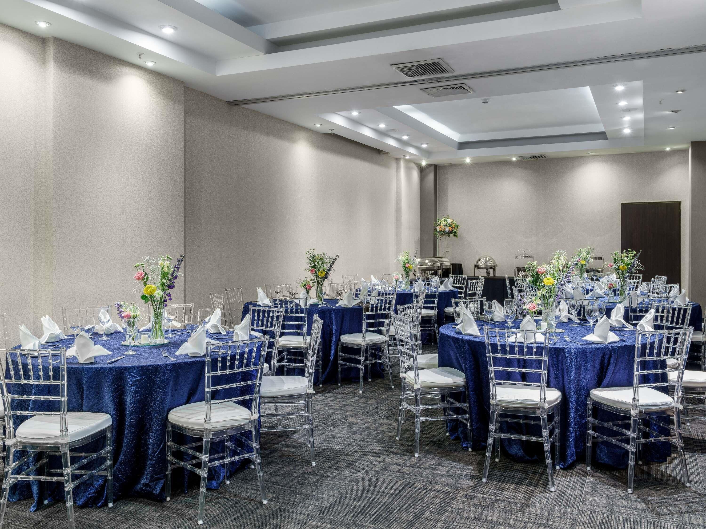 Un salón de conferencias con varias mesas, sillas transparentes y manteles azules.