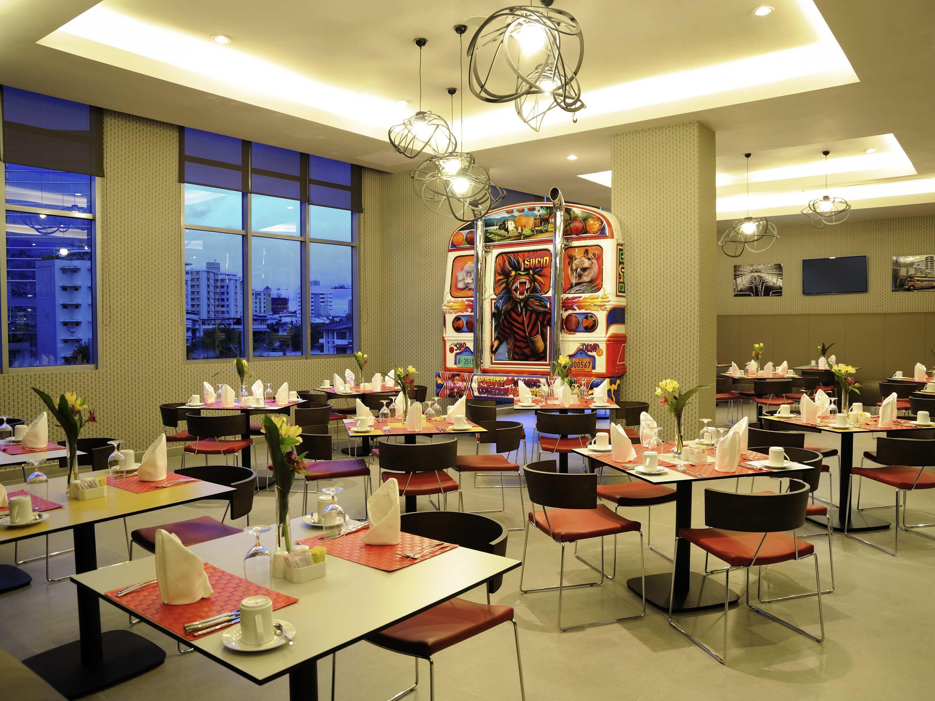 Une salle à manger d’hôtel avec des tables dressées, des chaises et une grande fenêtre donnant sur la ville.