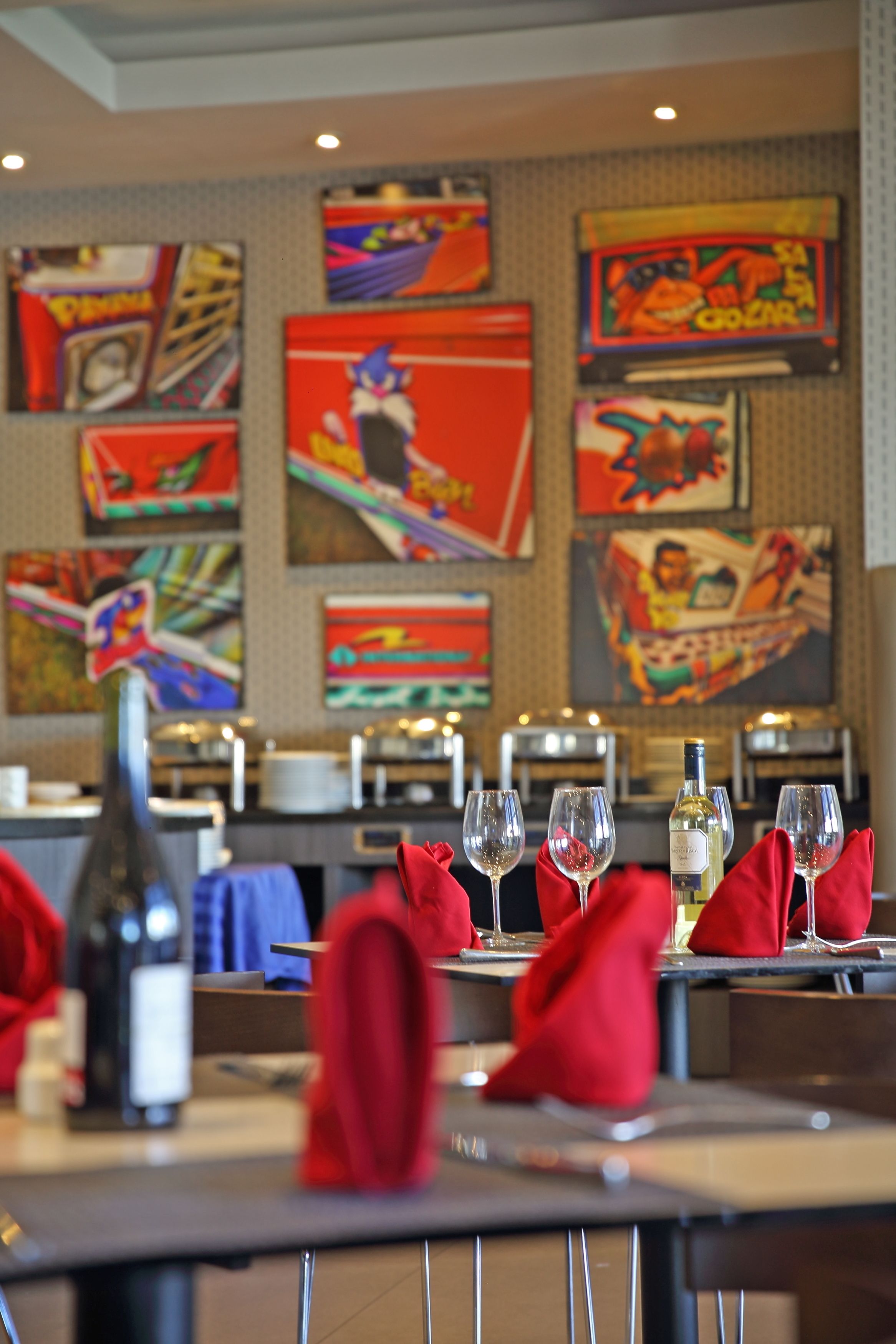 A restaurant interior with tables set for dining, featuring colorful artwork on the walls and a bottle of wine on the table.