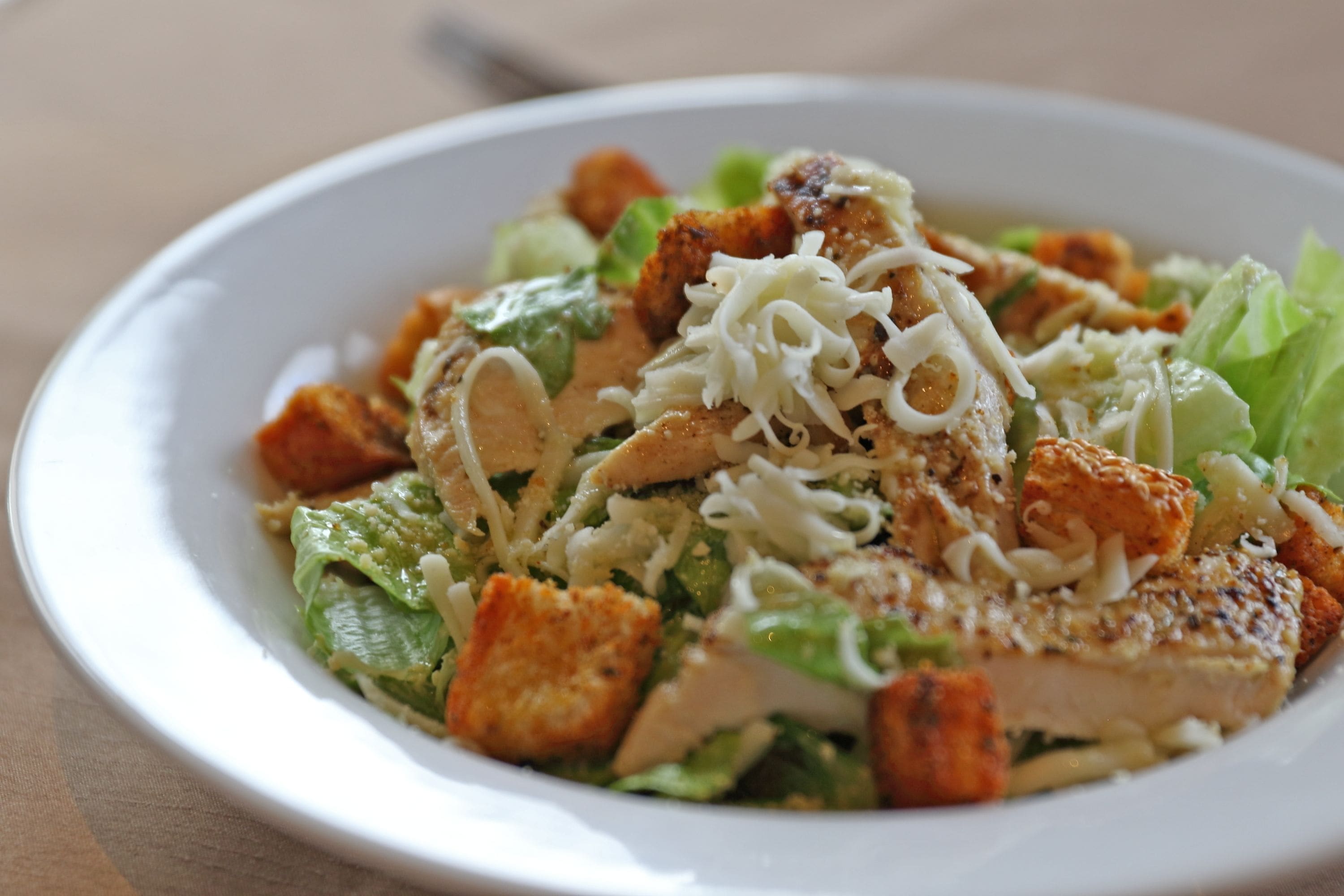 Um prato branco com uma salada de alface, croutons, queijo ralado e um pedaço de frango grelhado.