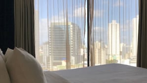 Een hotelkamer met een bed en uitzicht op een stadsgezicht door dunne gordijnen.