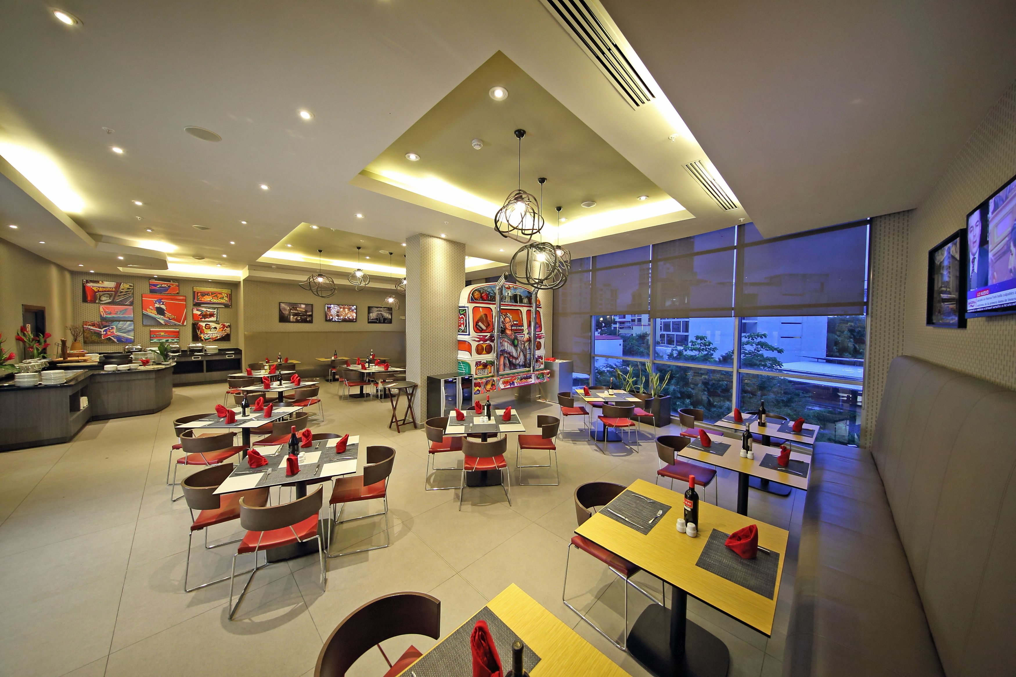 Un restaurante moderno con varias mesas y sillas, una zona de buffet central y grandes ventanales que ofrecen vistas al exterior.