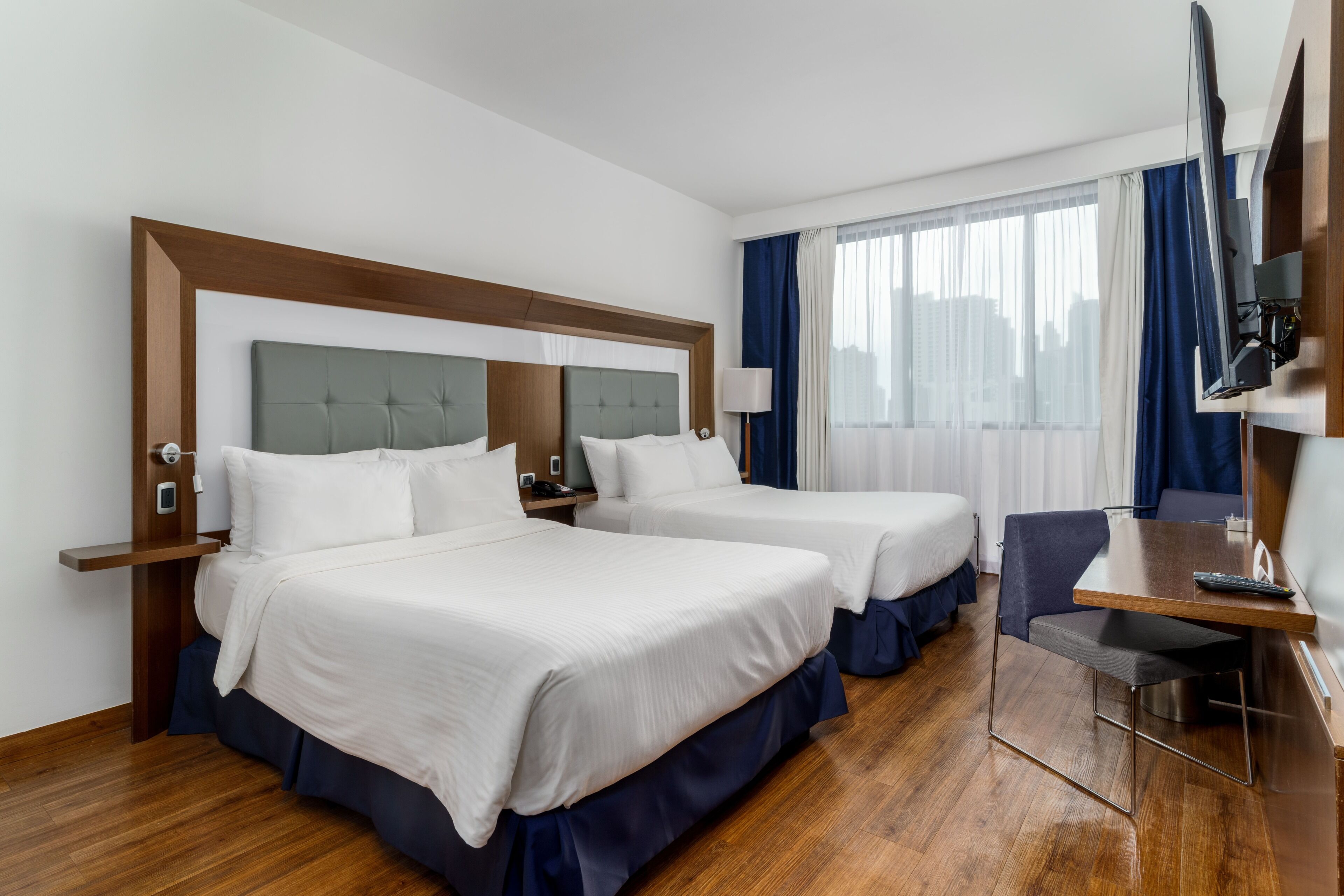 A hotel room with two beds, a desk, a chair, a TV, and a window with curtains.