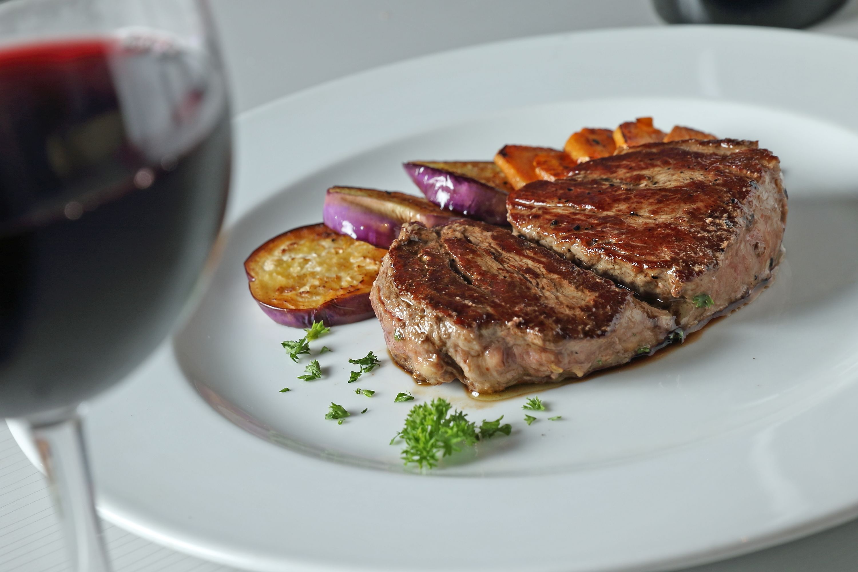 Une assiette comprenant un steak, des pommes de terre rôties et des oignons rouges, accompagnée d’un verre de vin rouge.