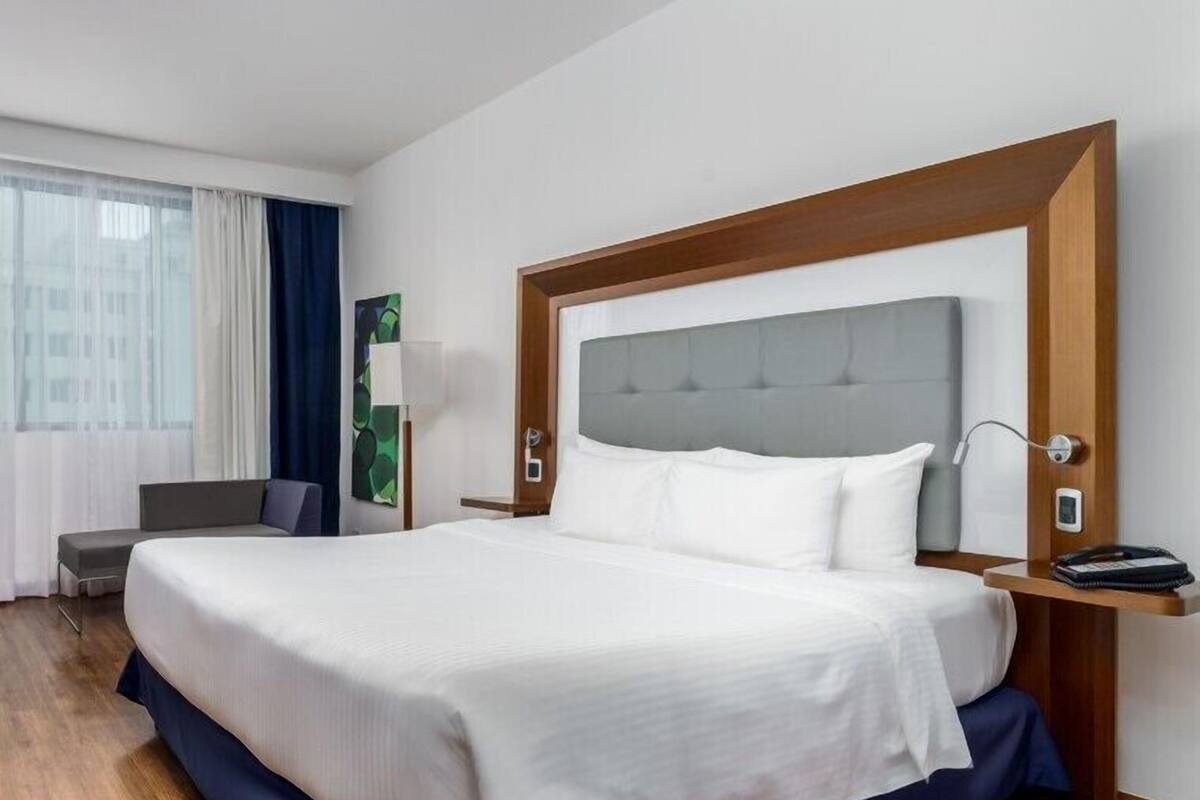 A hotel room with a large bed, a bedside table, a lamp, and a view of the city through the window.