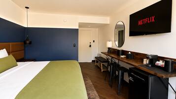 A hotel room with a bed, desk, chairs, a coffee maker, a television, and a mirror.