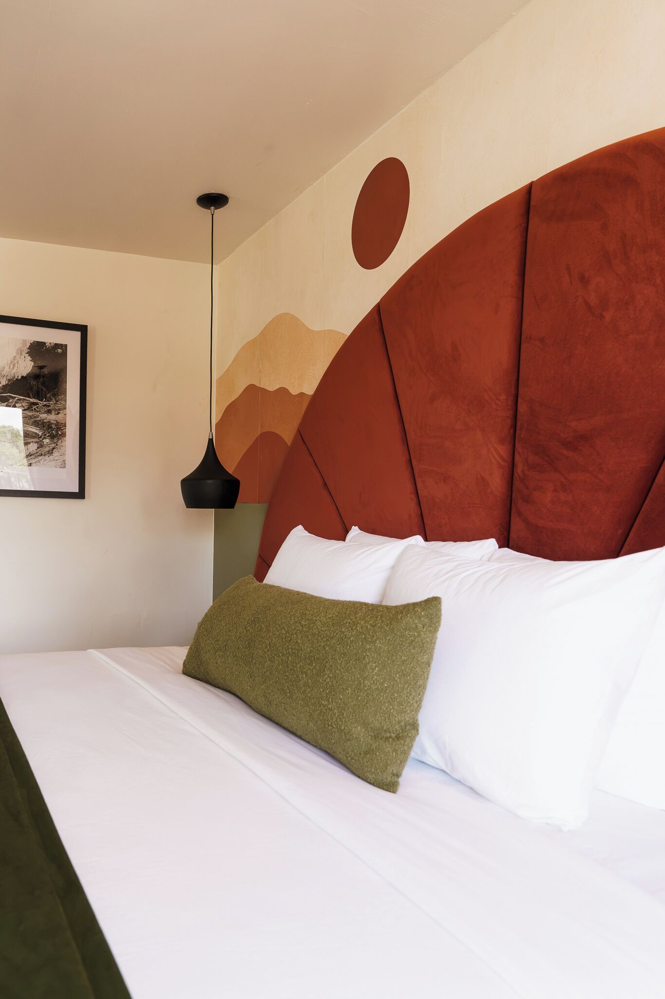 A bed with white bedding and a green pillow, a red headboard, a black pendant light, and a framed picture on the wall.