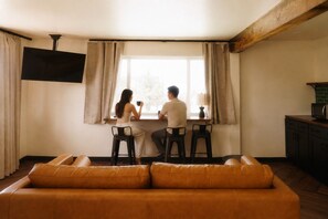 A living room with a sofa, a dining table with chairs, a TV mounted on the wall, and a window with curtains. - The Wesley Hotel (Page)