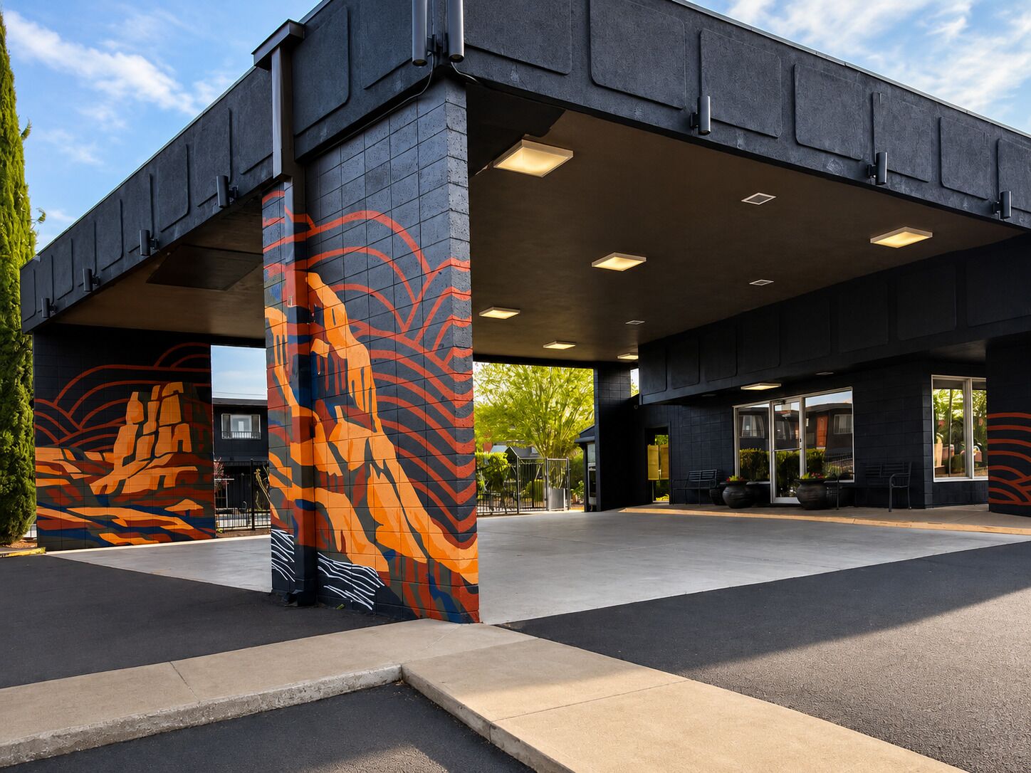 Un edificio moderno con un área de entrada cubierta, un mural en la pared y un espacio de descanso con plantas en macetas.