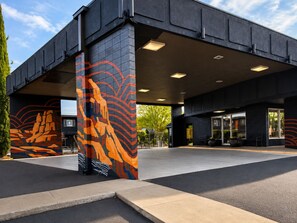 Un edificio moderno con un área de entrada cubierta, un mural en la pared y un espacio de descanso con plantas en macetas.