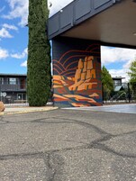 Un edificio con un mural debajo de un viaducto, un alto árbol de hoja perenne y una zona pavimentada con una valla.