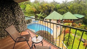 A wooden deck with two chairs and a table, a pool, and a view of a building with green roofs. - Tabulia Tree Hotel and Villas (Quepos)
