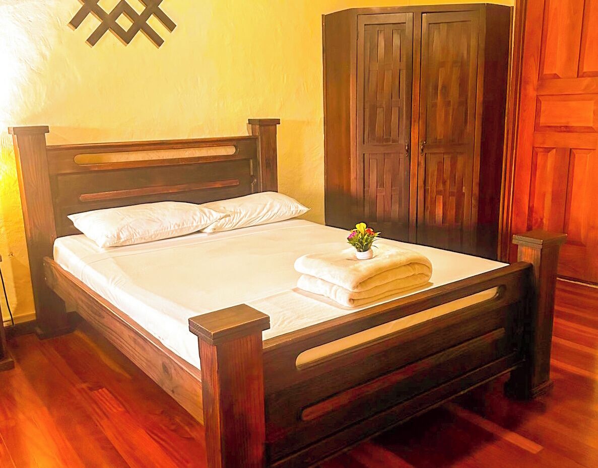 A wooden bed with white linens, a nightstand, a wooden wardrobe, and a wall-mounted light fixture.