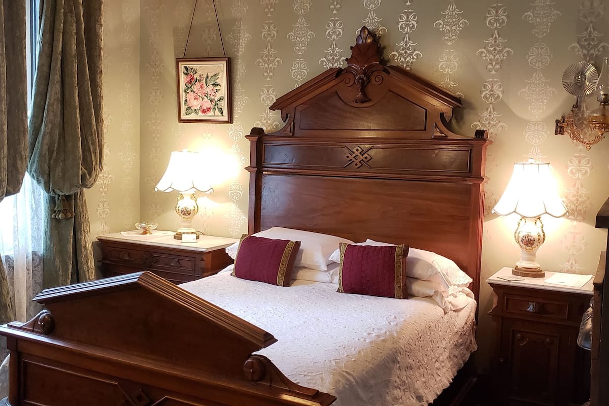Un dormitorio tradicional con cabecera de madera, dos mesitas de noche con lámparas, un radiador y una ventana con cortinas.