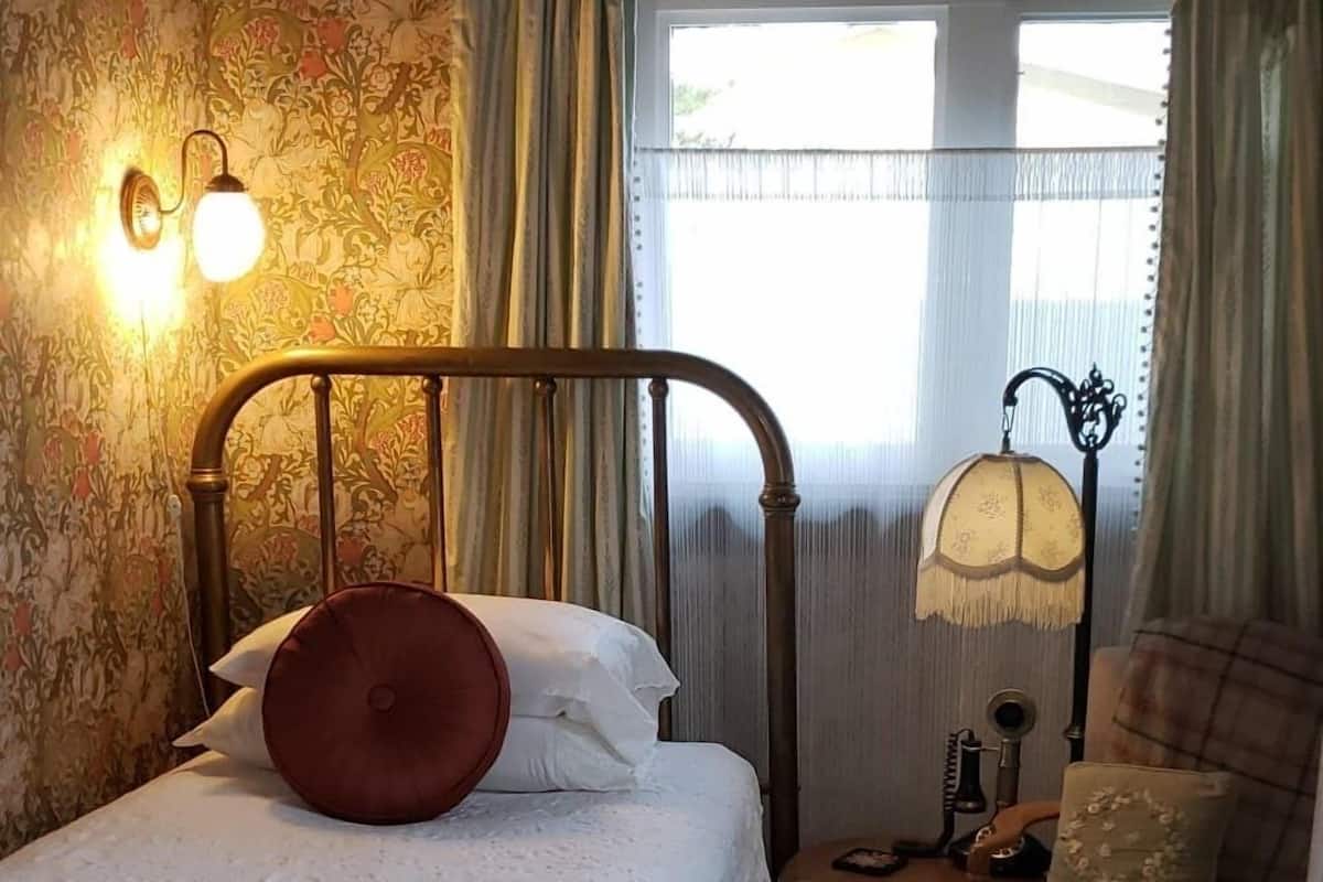 Un dormitorio con cama, mesita de noche, una silla, radiador y ventana con cortinas.