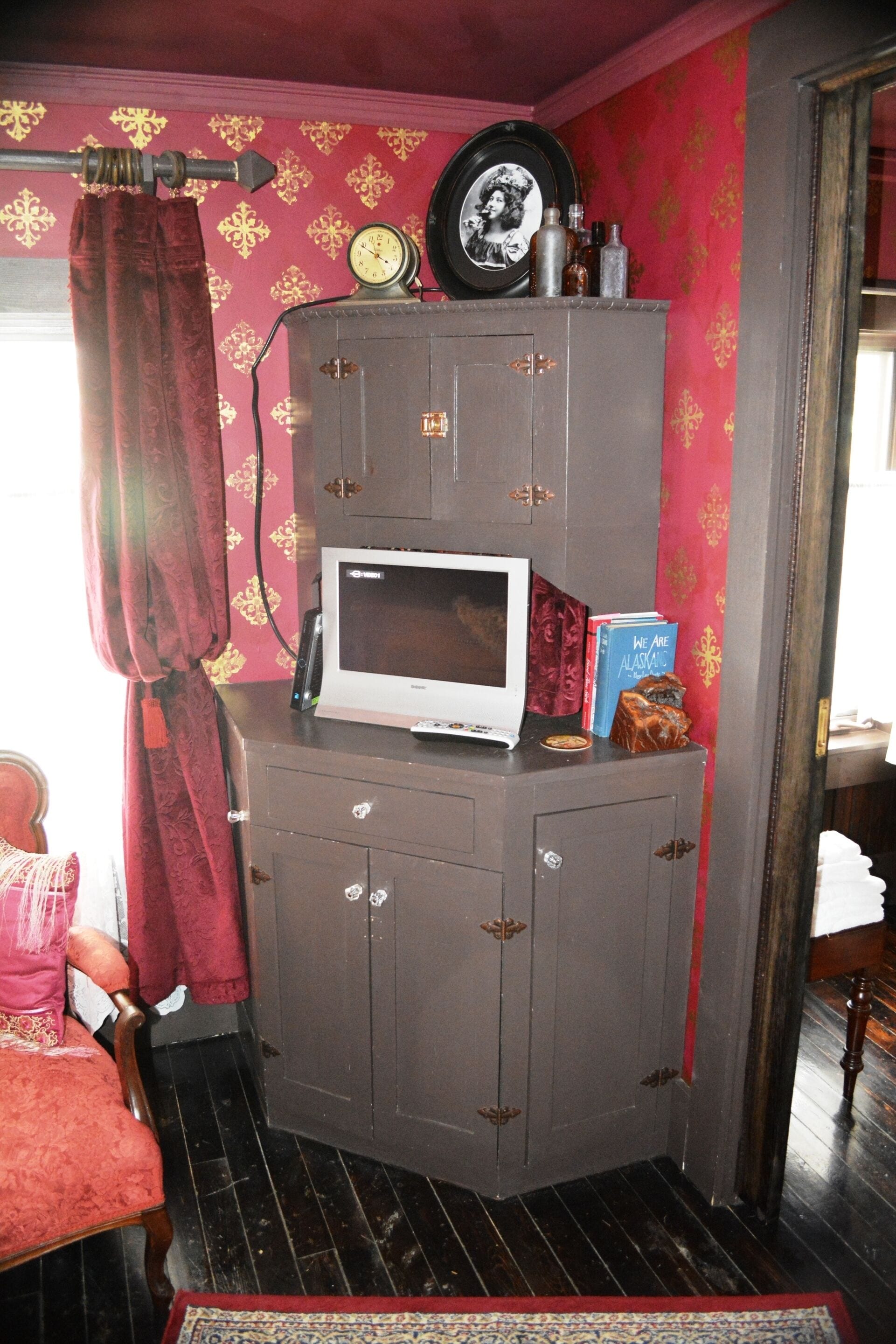 A corner room with a vintage-style cabinet, a microwave, a clock, and a framed photo on the wall.