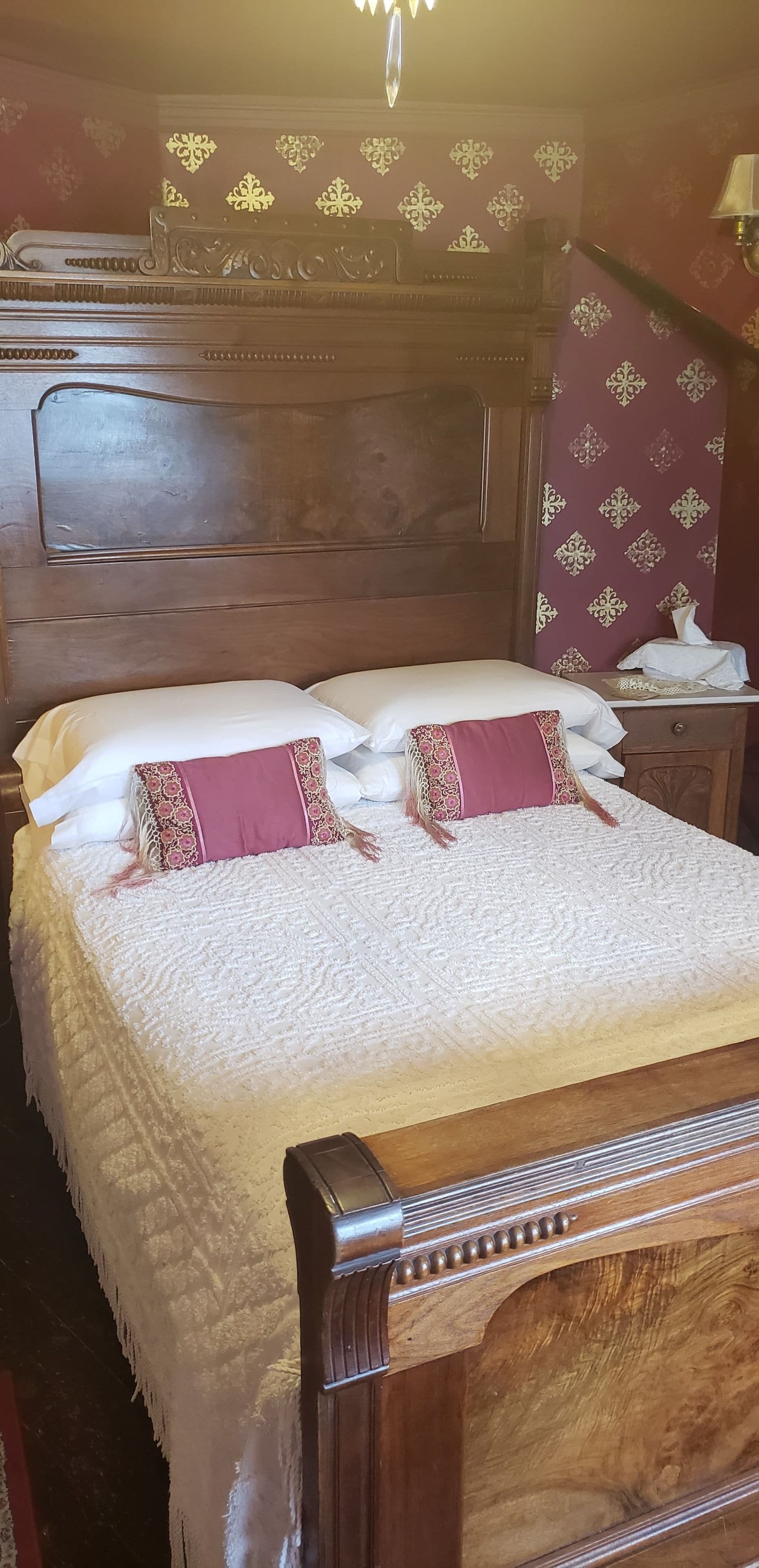 A neatly made bed with a white quilt and two decorative pillows, a wooden headboard with intricate designs, and a patterned wall in the background.