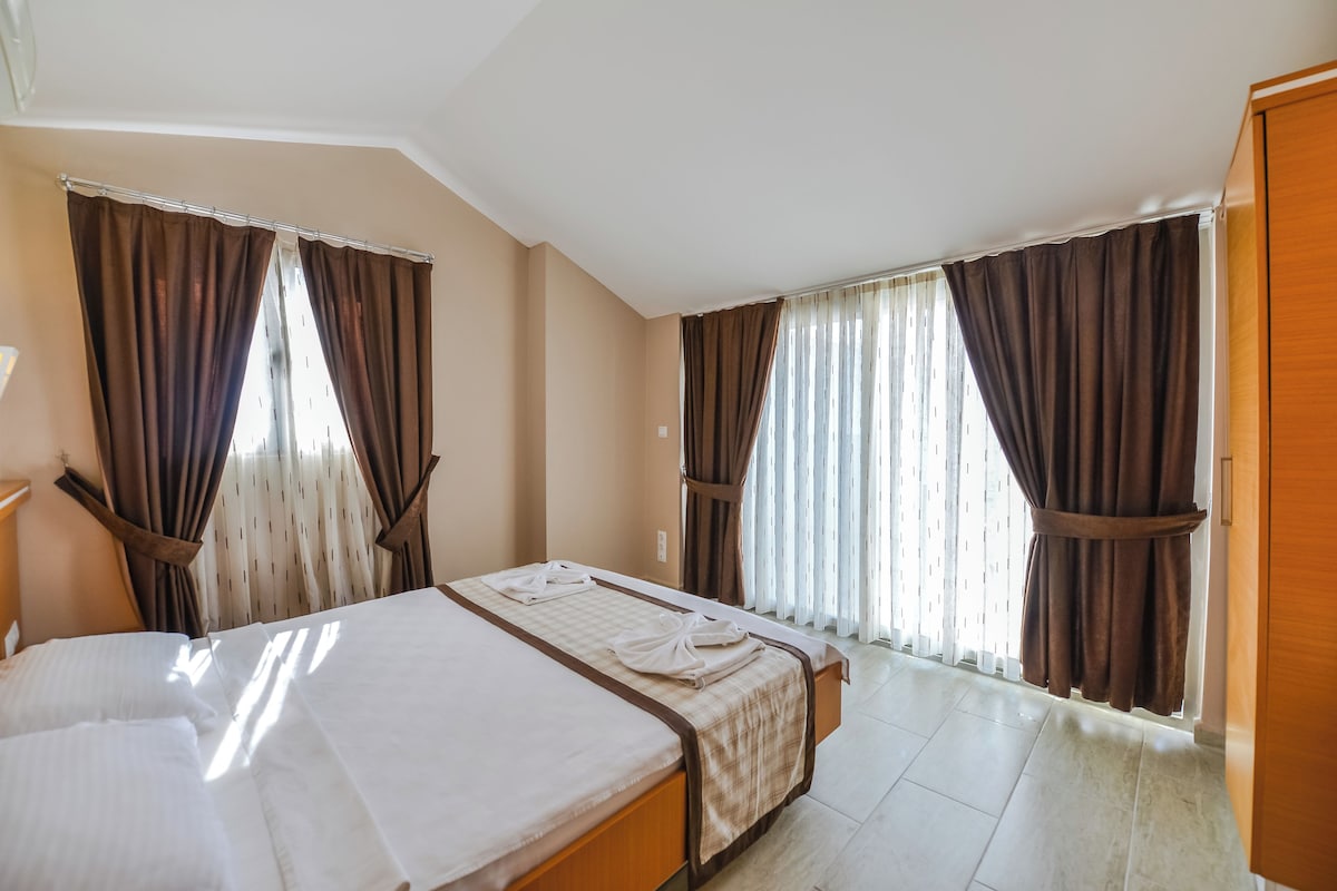 A hotel room with a bed, a desk, and two windows with curtains.
