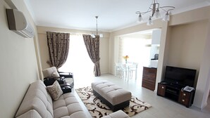 A modern living room with a beige sofa, a small ottoman, a TV stand with a flat-screen TV, and a dining area with a table and chairs. - Diana Residence (Fethiye)