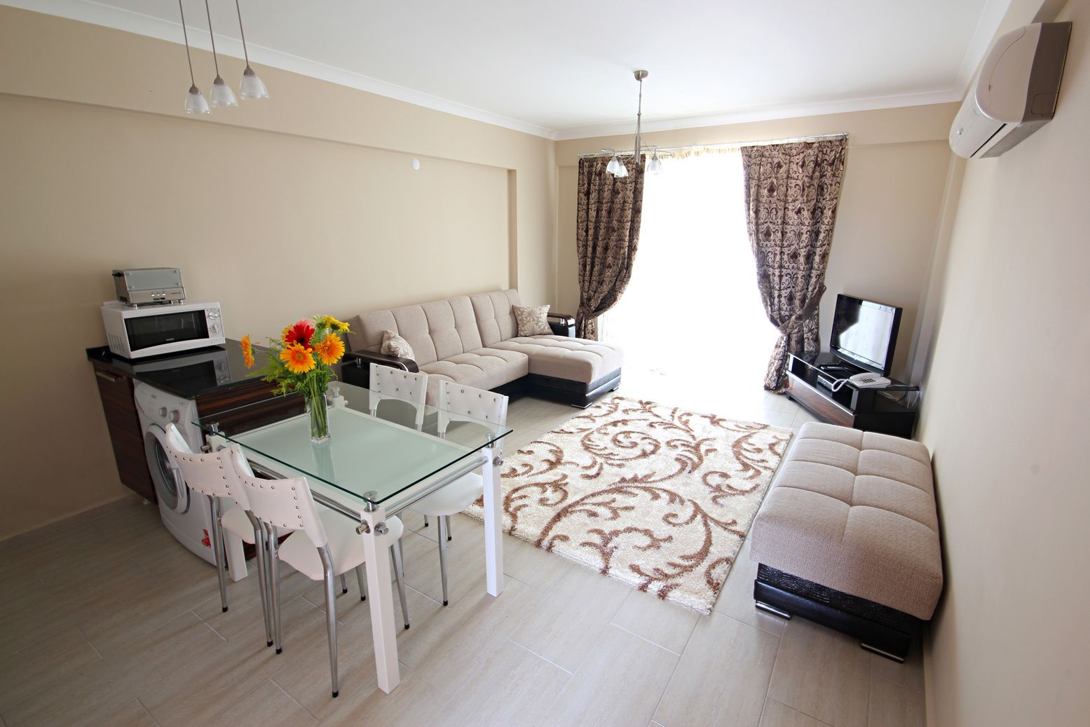 Una sala moderna con una mesa de comedor de vidrio, sillas blancas, un sofá beige, un televisor y una lavadora.