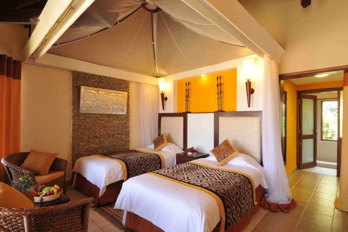 A hotel room with two beds, a wicker chair, a fruit bowl, and a brick wall.