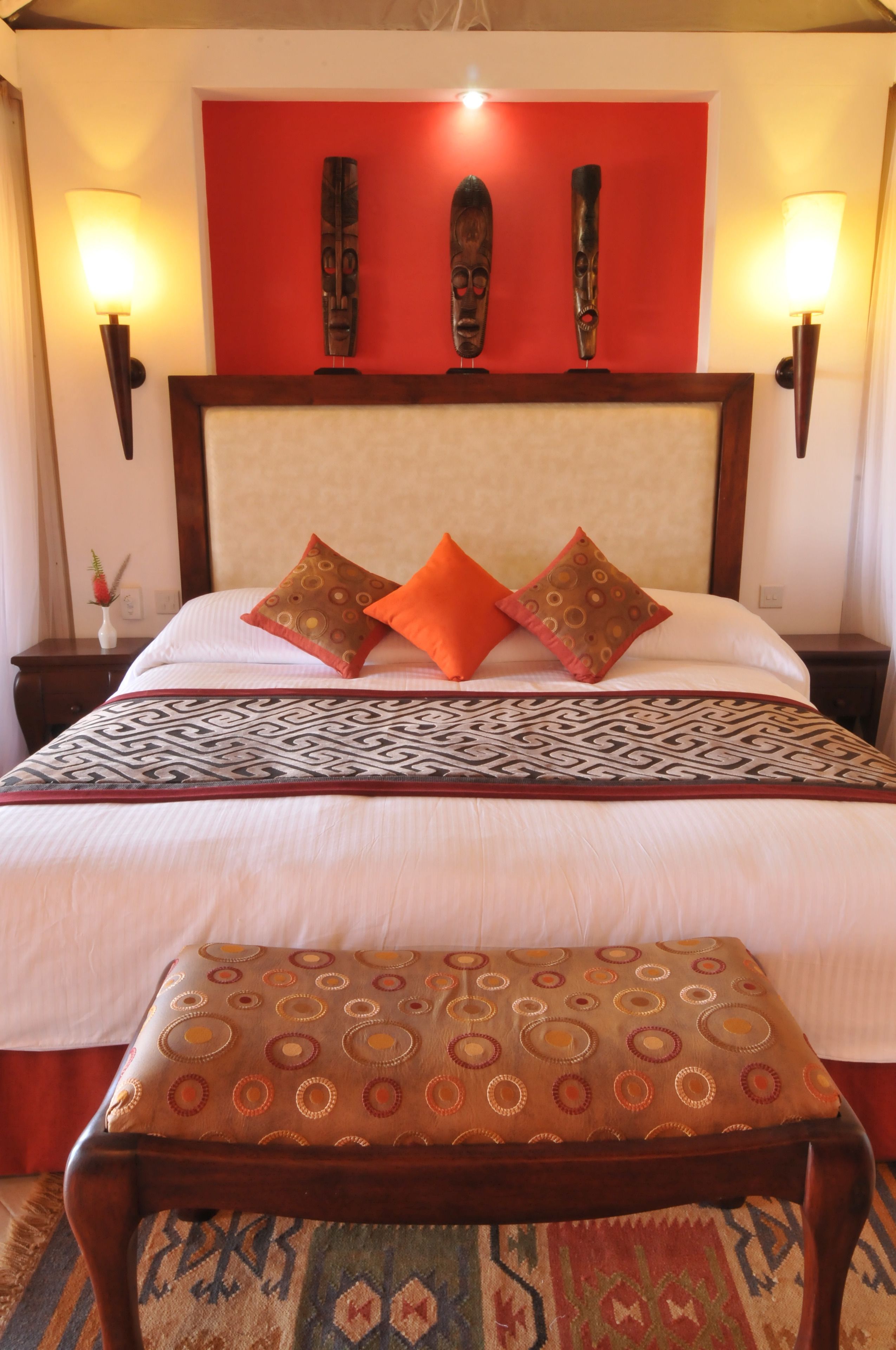 Una habitación de hotel con una cama grande, dos lámparas fijadas a la pared, una pared de acento roja y una manta decorativa estampada.