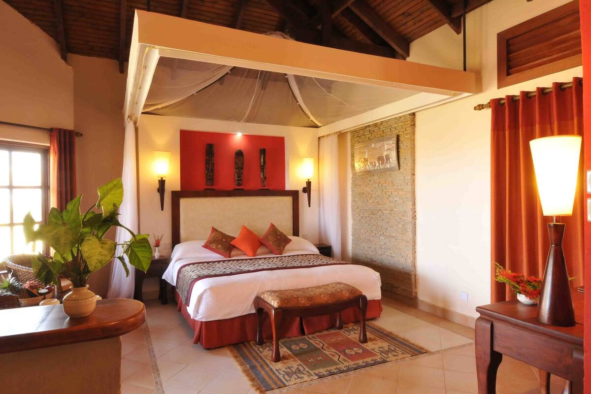 A bedroom with a bed, bedside tables, a nightstand, a window, a plant, and a wall-mounted lamp.