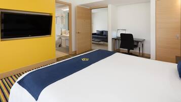 Quarto de hotel com cama, escrivaninha, cadeira, televisão e banheiro visível por uma porta aberta.