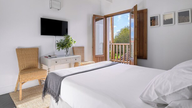 Une chambre avec un lit, un téléviseur fixé au mur, une porte en bois donnant sur un balcon et un fauteuil en osier.