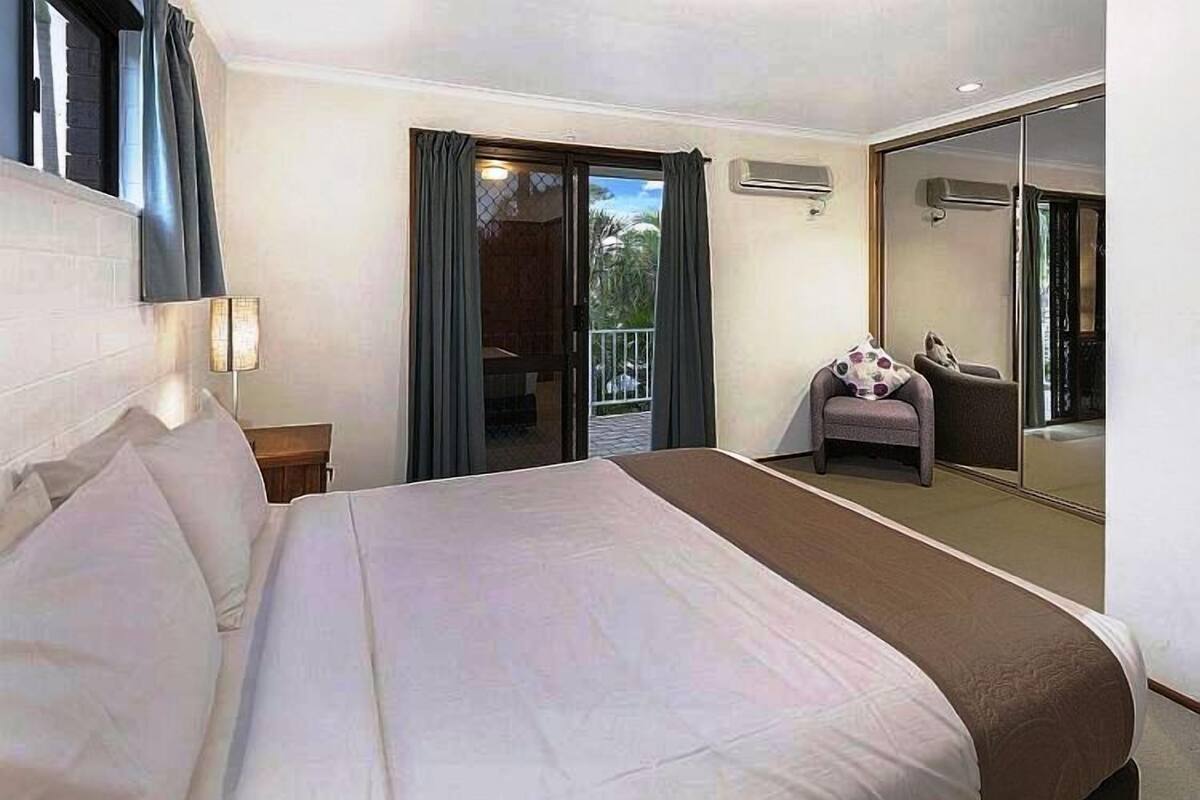A hotel room with a large bed, a small seating area with a chair and a table, a balcony with a view, and a sliding glass door.